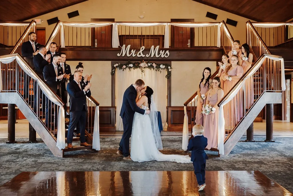 A wedding ceremony with bride and groom kissing, surrounded by bridal party and guests on a staircase decorated with white drapery and flowers, and a sign that reads 'Mr & Mrs' in the background.