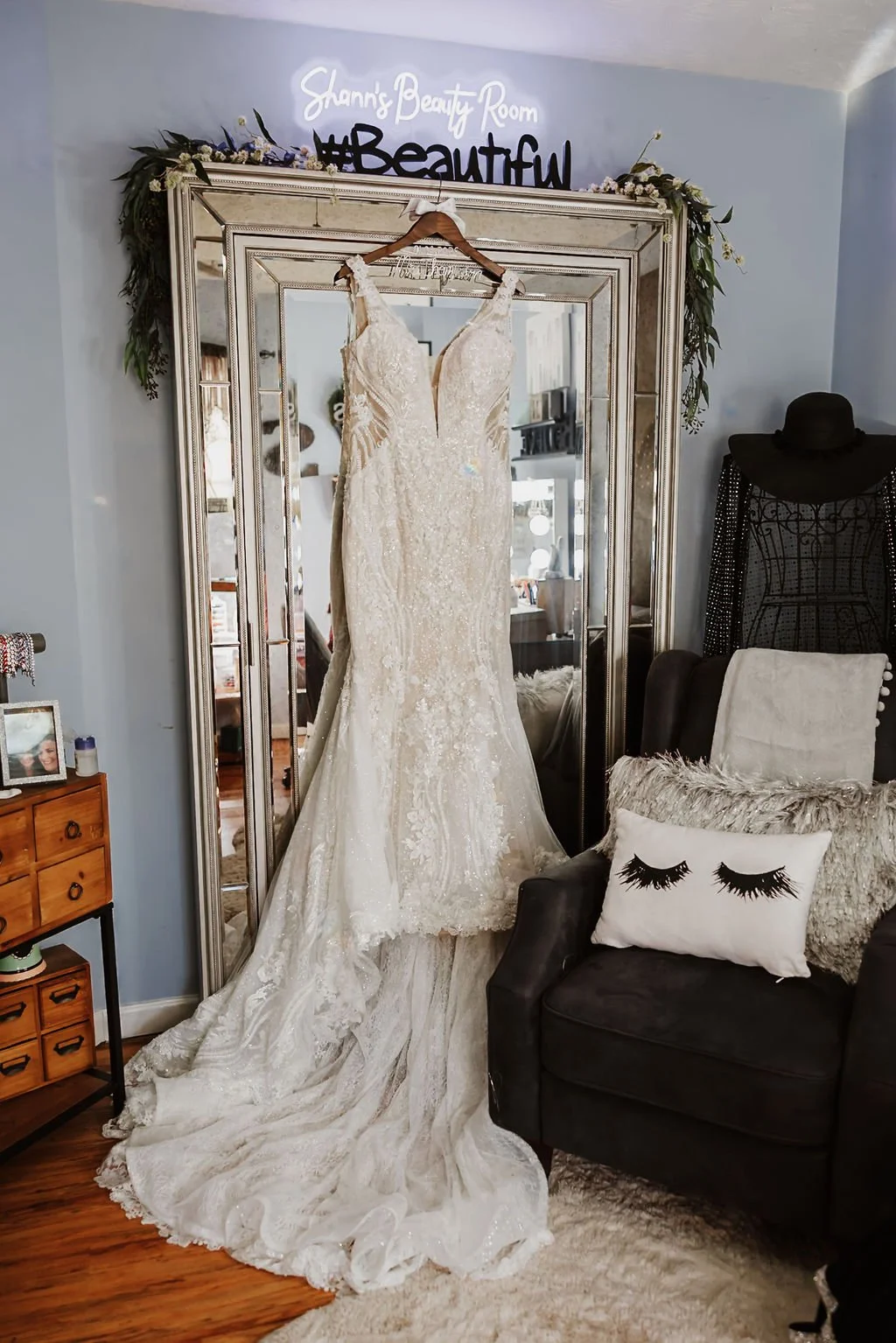 A wedding dress hanging on a wooden hanger in front of a large decorative mirror with a sign above that reads 'Shann's Beauty Room #Beautiful.' The dress is cream-colored with intricate lace details and a long train. Oscar Event Center Fairfield, Oh