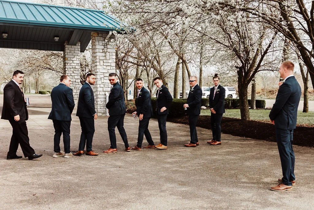 Nine men in tuxedos standing in a line outdoors, playing a game of dominoes, with a stone building and trees in the background.