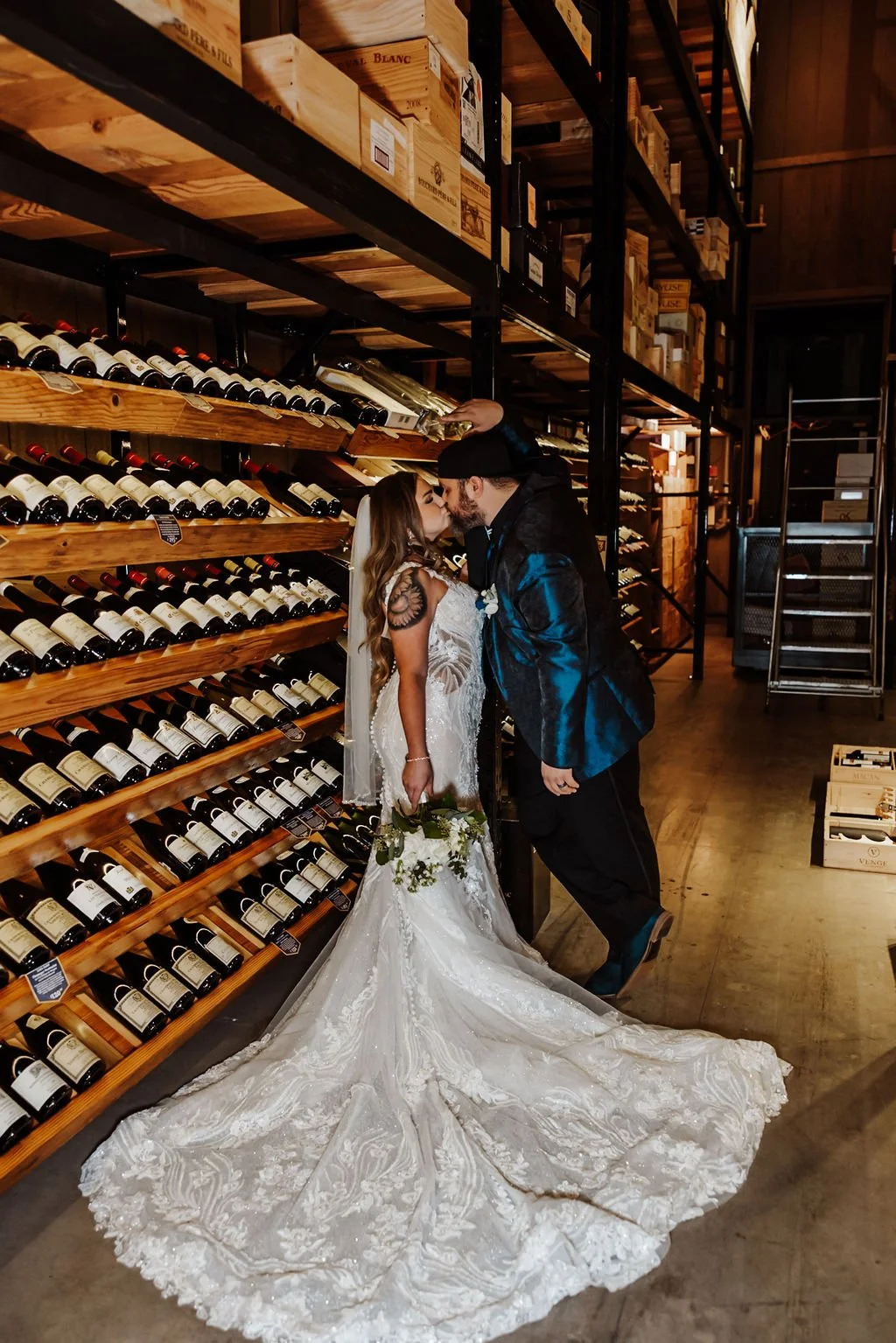 A bride and groom kissing in a wine store, with wine bottles on wooden shelves behind them.