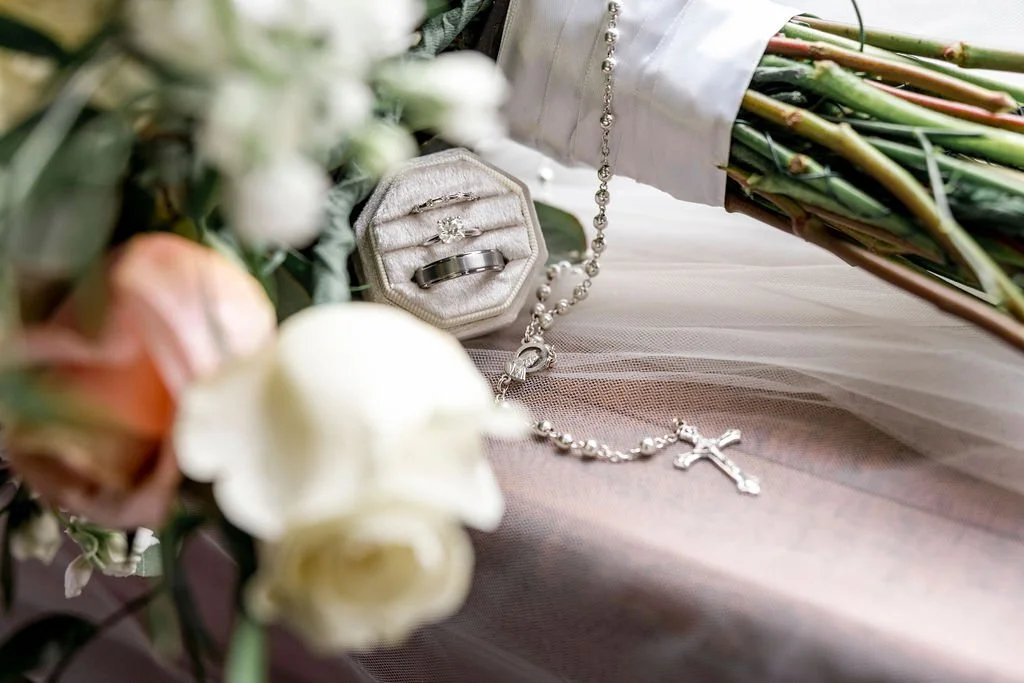 A bouquet of flowers, wedding rings in a box, and a rosary with a cross on a white fabric surface.