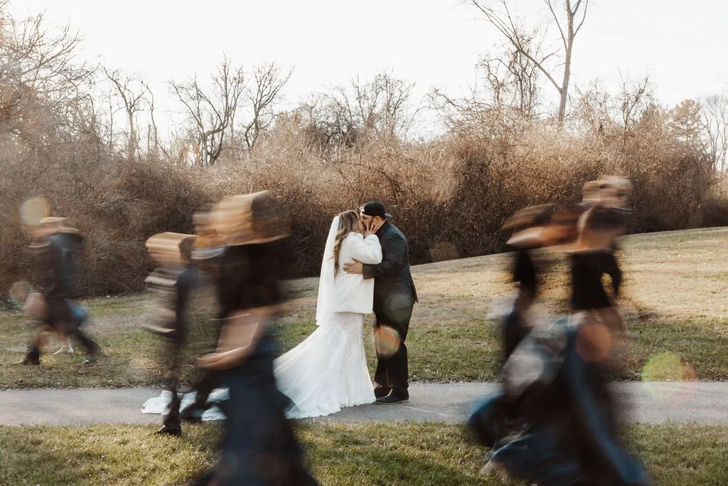 A bride and groom sharing a kiss on a walking path with blurred wedding guests passing by in motion in the foreground, outdoor setting with trees and bushes in the background.