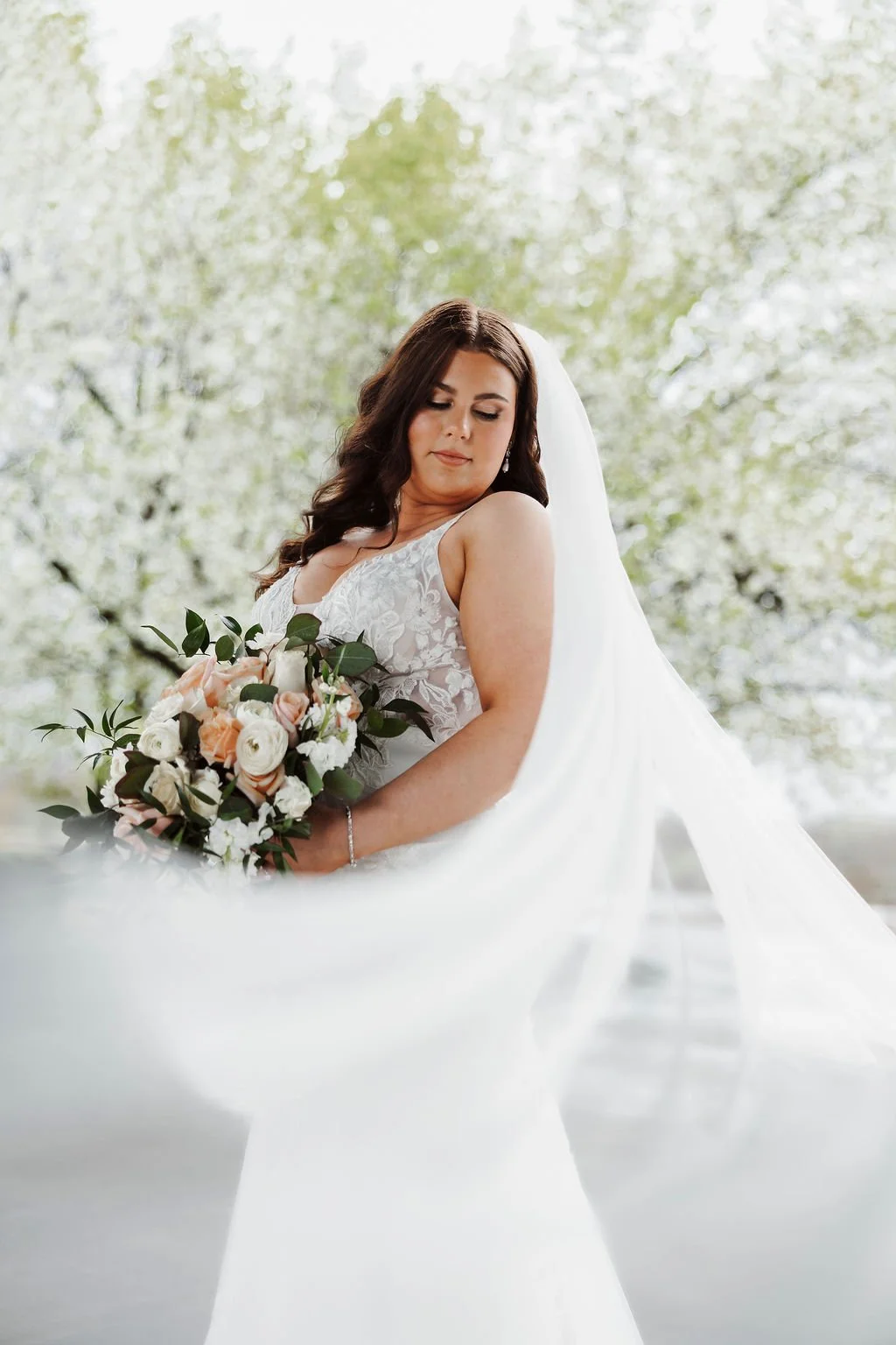 A bride in a white wedding dress holding a bouquet of flowers, standing outdoors in front of trees with spring foliage.