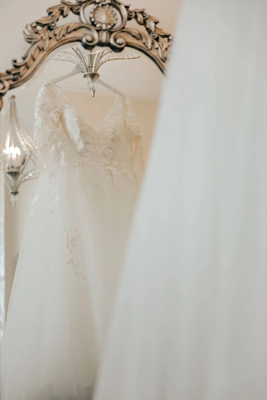 A wedding dress hanging from a decorative ceiling light fixture, reflected in a mirror with an ornate frame.