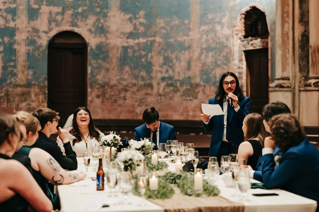 People at a wedding reception, sitting at a long table, listening to a man with glasses and long hair in a blue suit giving a speech, with candles and floral decorations on the table, in an ornate, historic venue.