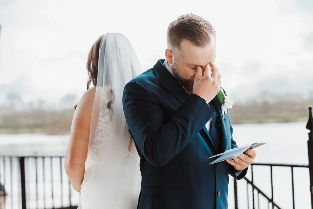Groom reading a letter or note during a wedding ceremony, with bride standing behind him and wearing a veil, outdoors near water.