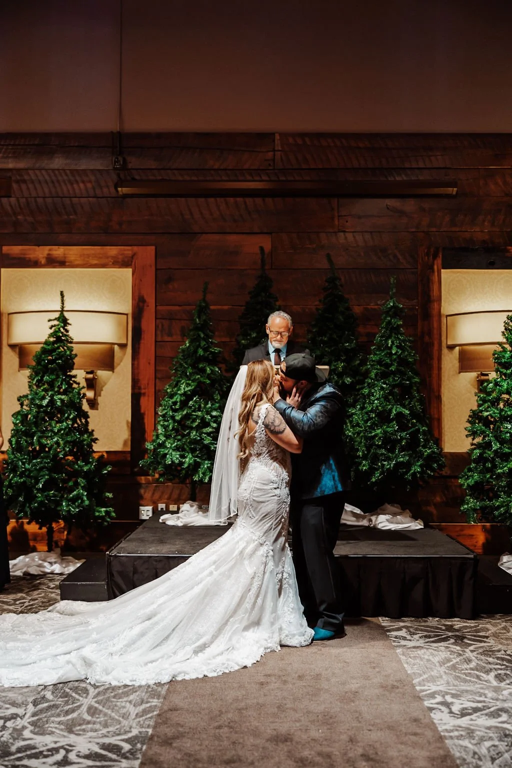 A wedding ceremony with a bride and groom kissing in front of an officiant, surrounded by Christmas trees and warm lighting.