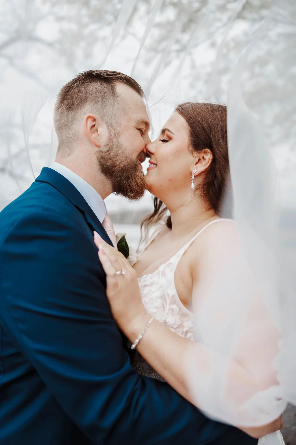 A bride and groom are nose-to-nose, smiling and embracing, with a soft veil surrounding them in an outdoor setting.