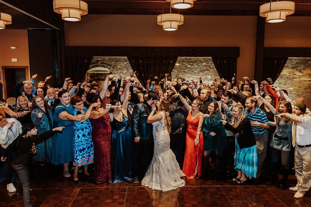 Group of people celebrating a wedding reception, with the bride and groom in the center, dancing under warm lighting in a decorated indoor venue.