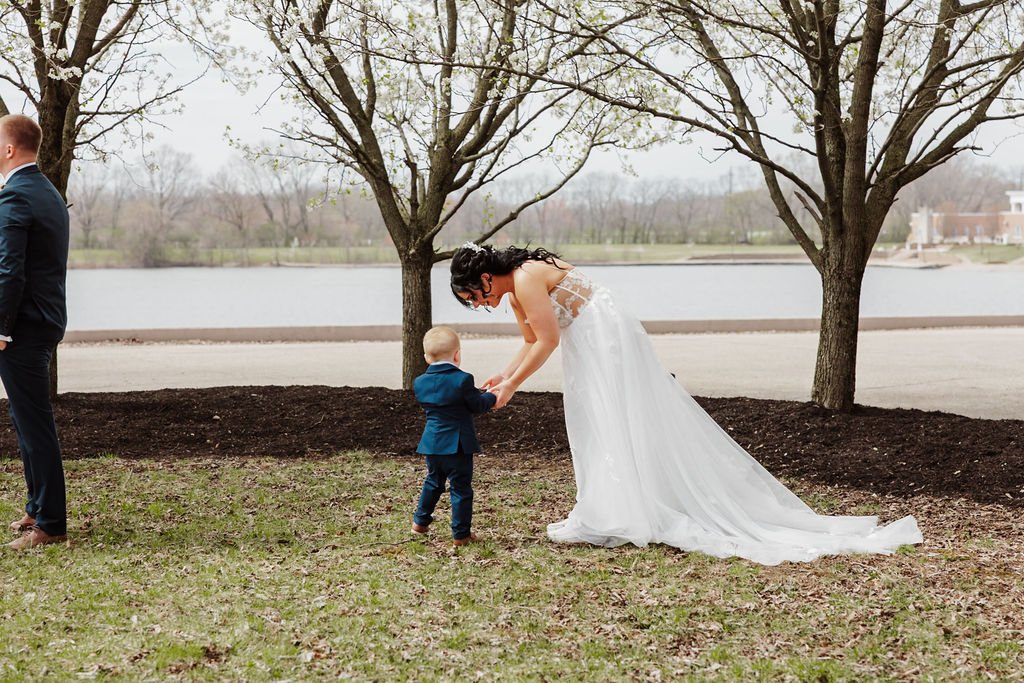 A woman in a white wedding dress bending down and holding hands with a young boy in a suit outside near a lake, with trees and a cloudy sky in the background.
