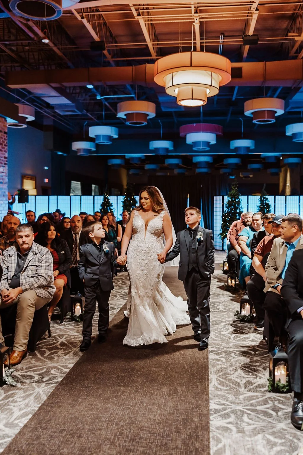 A bride holding hands with two young boys as they walk down the aisle during a wedding ceremony in a decorated indoor venue with guests seated on both sides and Christmas trees in the background.