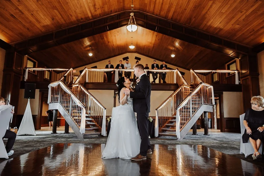 A bride and groom dance together at their wedding reception inside a wood-paneled hall with a high vaulted ceiling. The couple is centered on the dance floor with guests seated around them, and a band is visible on the upper balcony in the background