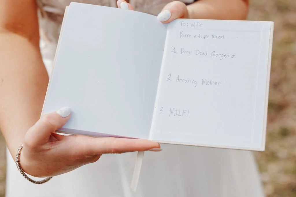Open small white notebook with handwritten list on right page, held by person with light skin and white-painted nails, outdoors.