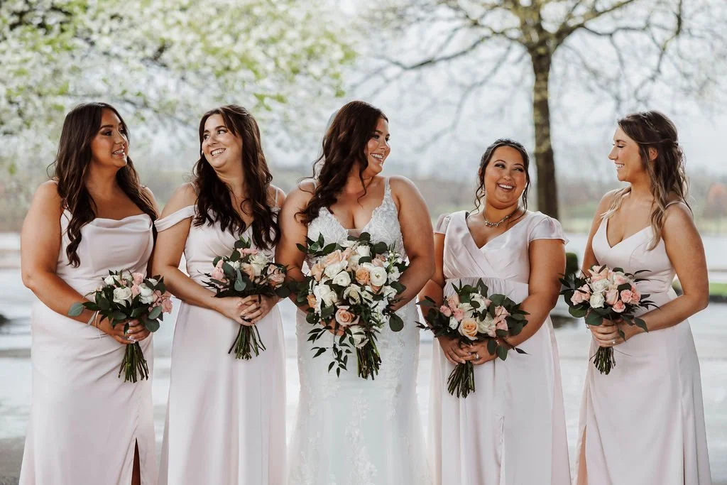 Bridal party with women in white dresses holding bouquets, standing outdoors with trees and a lake in the background.
