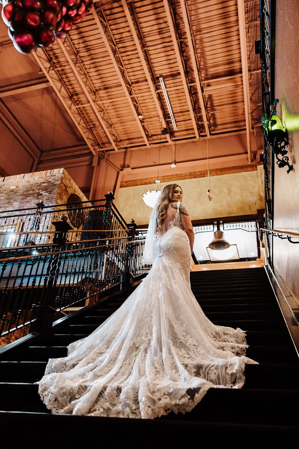 A bride in a lace wedding gown with a train, standing on a staircase in an indoor venue with exposed wooden beams and brick accents.