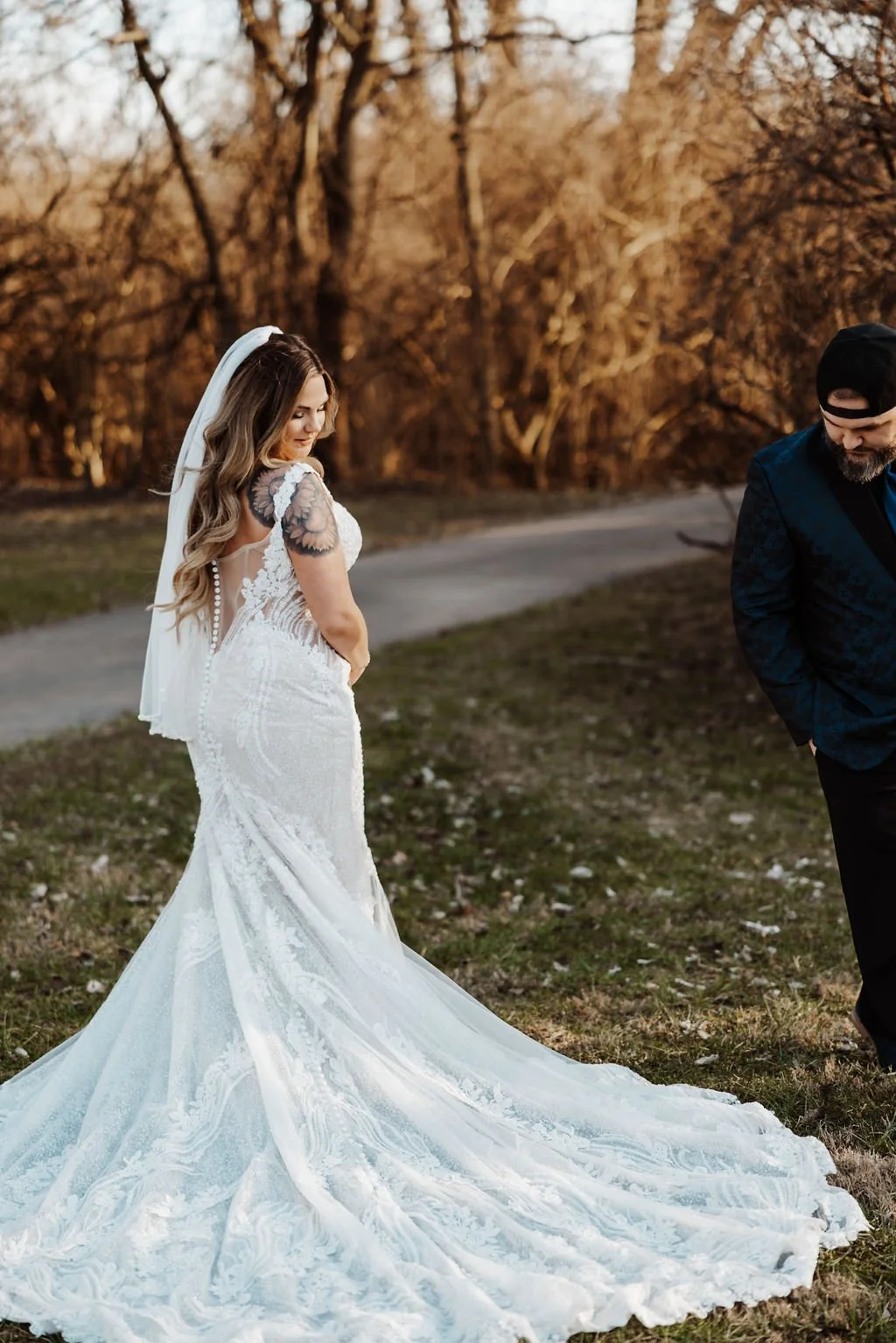 A bride in a white wedding dress with lace details and a long train, standing outdoors on grass with trees in the background, smiling and looking down, with a man in a dark jacket and cap partially visible beside her.