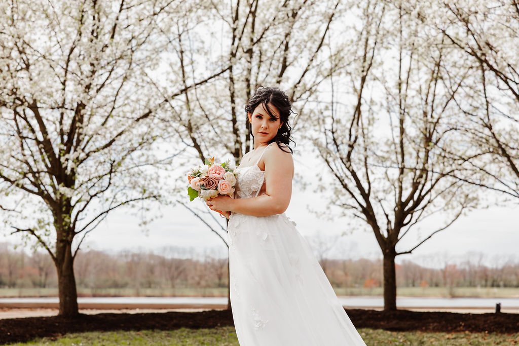 A bride in a white wedding dress holding a bouquet of pink and white roses outdoor with trees and a cloudy sky in the background.