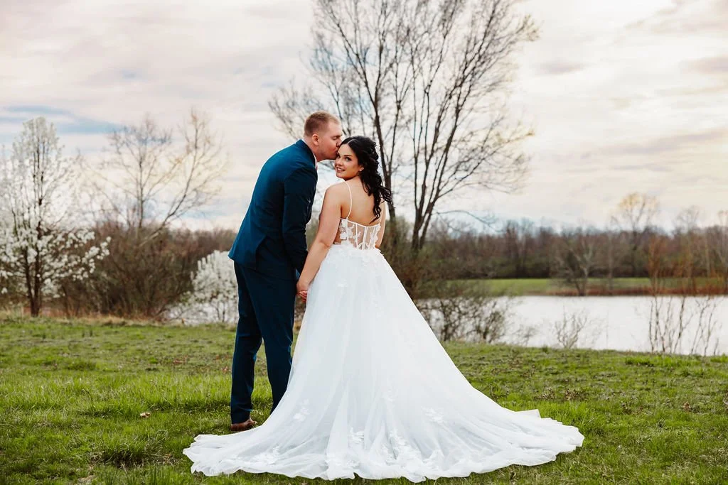 A bride and groom standing on grass near a lake, with the groom kissing the bride on the cheek, in a scenic outdoor setting during daytime.