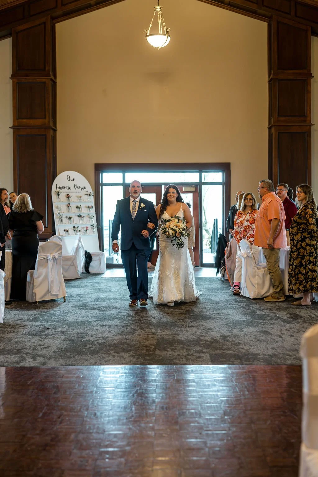 A bride walking down the aisle accompanied by a man in a blue suit at a wedding ceremony in a spacious hall with high ceilings and wooden paneling.