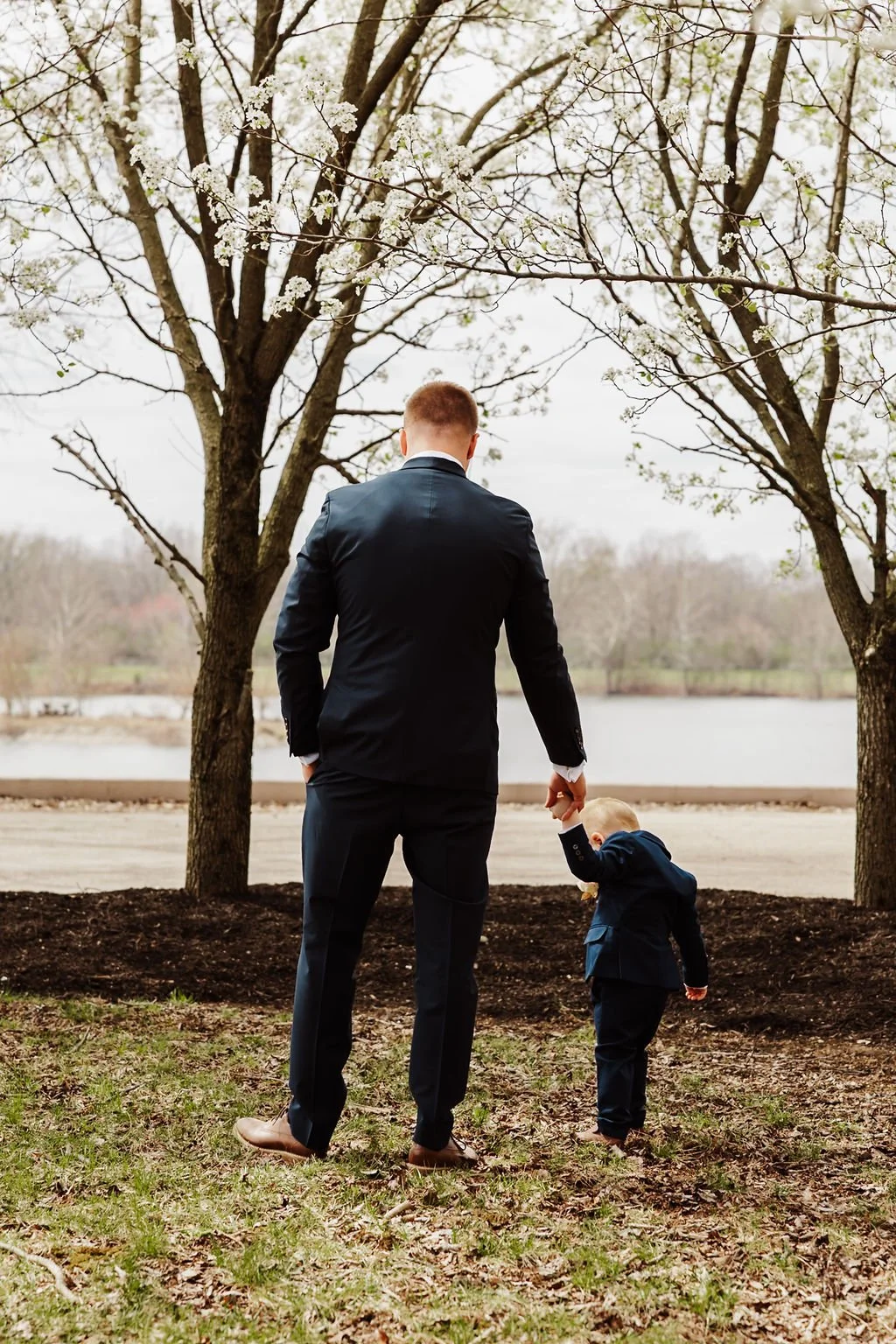 A man and a child walking outdoors near a lake, with the man holding the child's hand. Both are dressed in dark suits. The scene is framed by trees with white blossoms, indicating early spring.