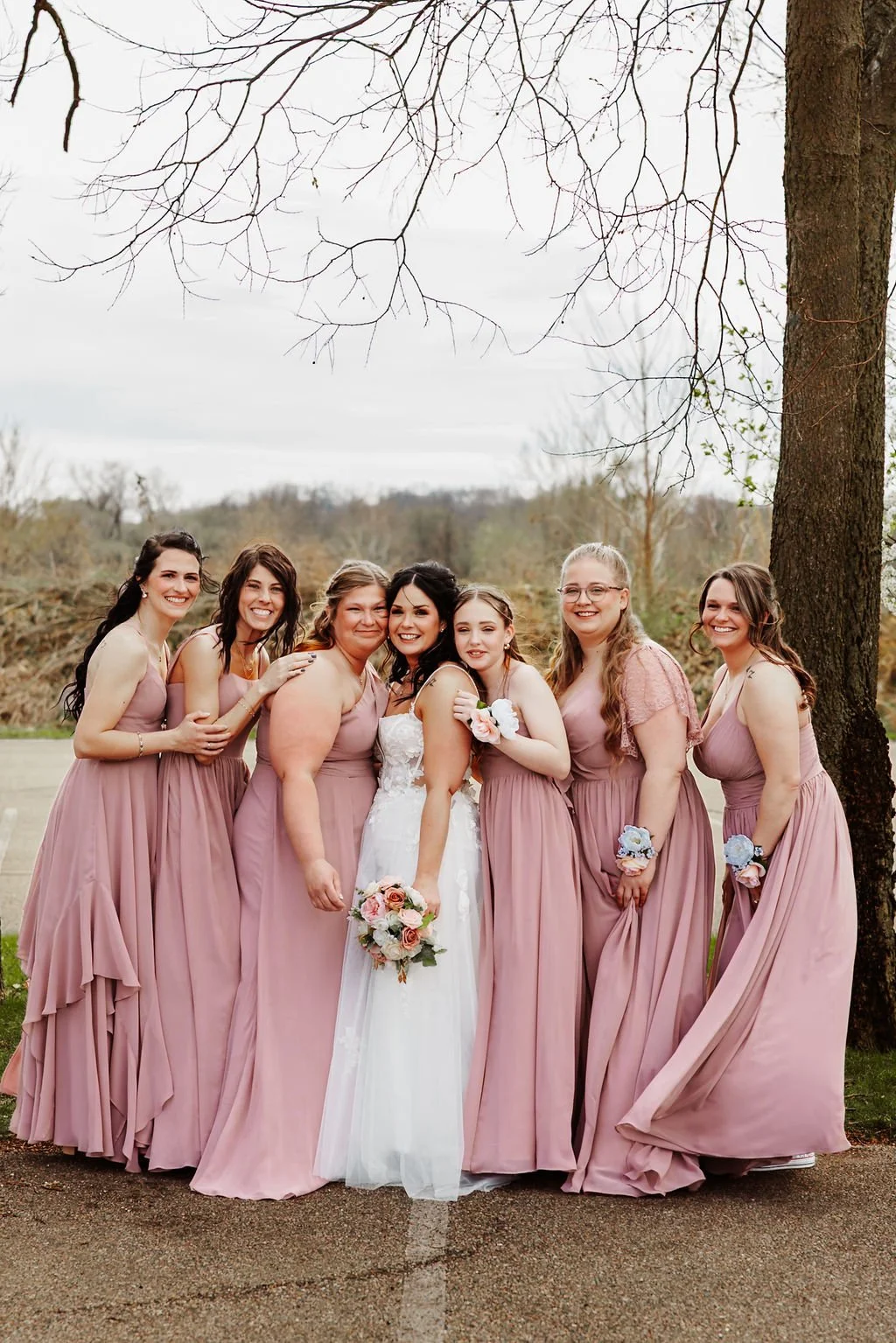 A group of seven women, centered around a bride in a white wedding dress, standing outdoors on a cloudy day. The women are dressed in matching mauve dresses, and some are holding bouquets. They are smiling and hugging, with trees and a cloudy sky in 
