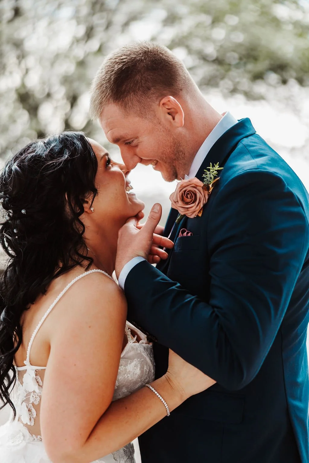 A bride and groom close together, smiling and touching noses, on their wedding day outdoors.