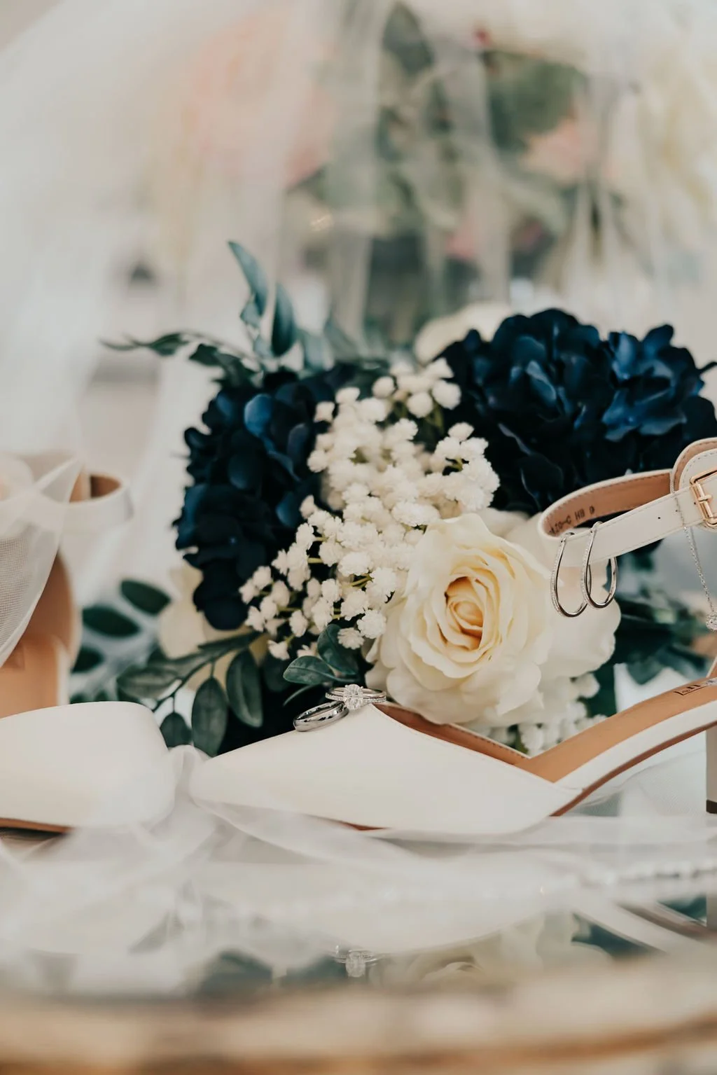 Wedding bouquet with white and navy flowers, white high heel shoe with a ring, wedding rings, and jewelry on a table with a mirror reflection.