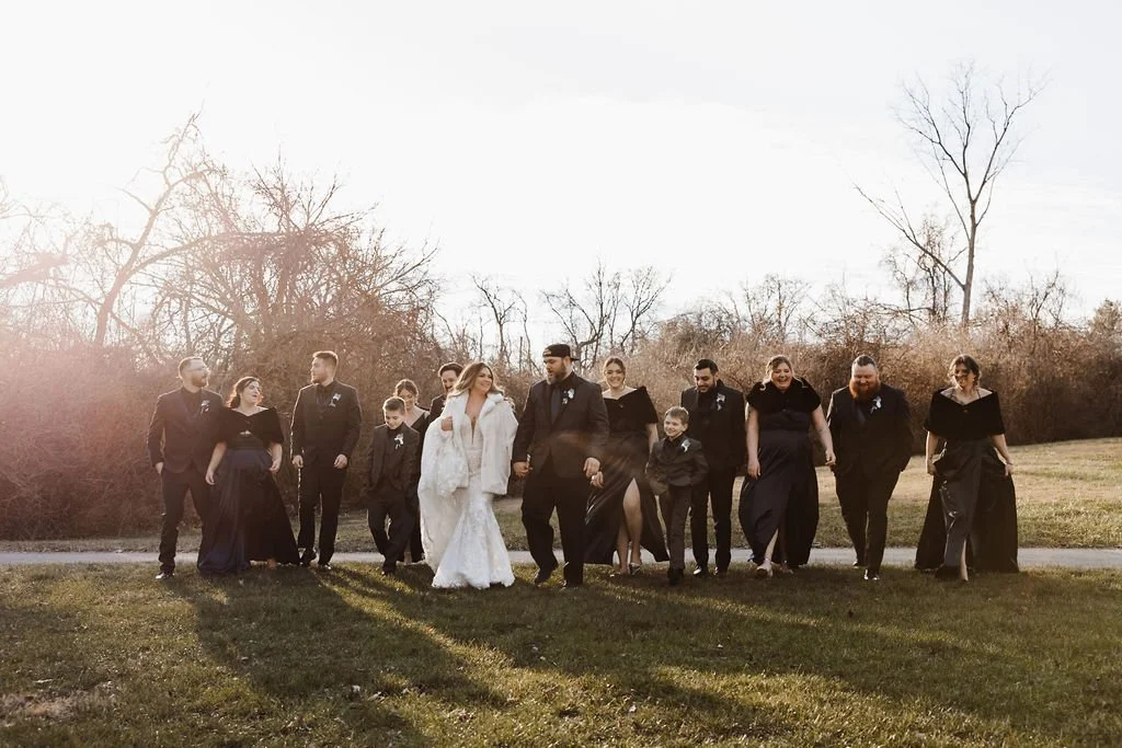 A wedding party walking outdoors on a grassy area with leafless trees in the background during late afternoon.