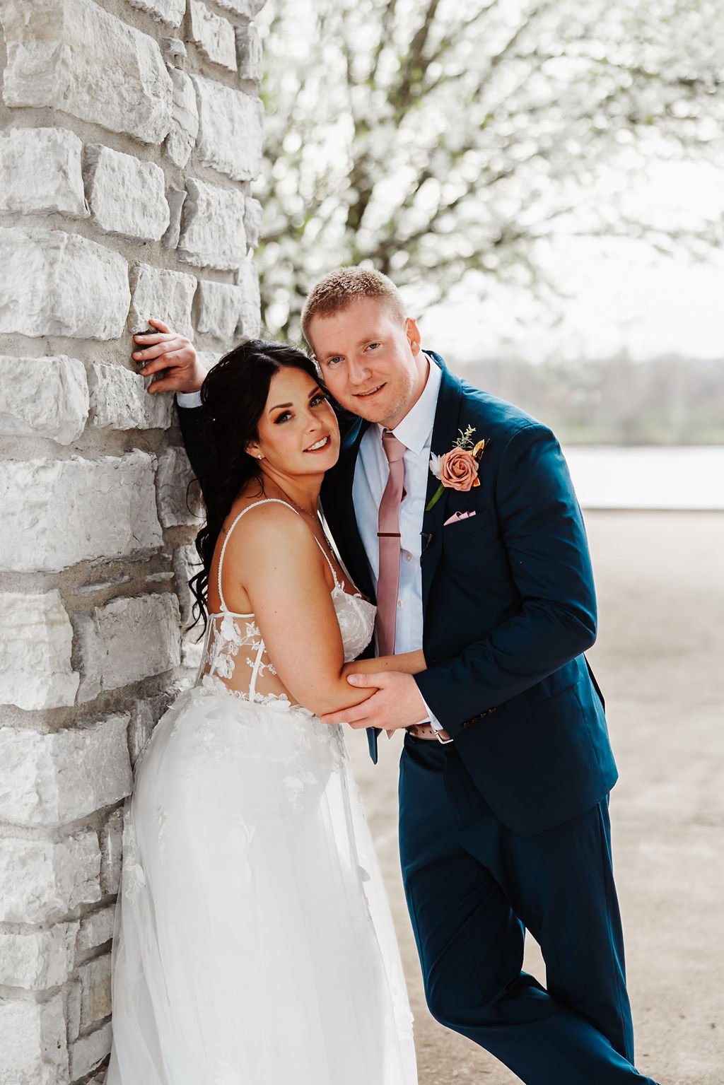 A newlywed couple in wedding attire, posed outdoors near a stone wall with a blurred background of trees and a body of water.