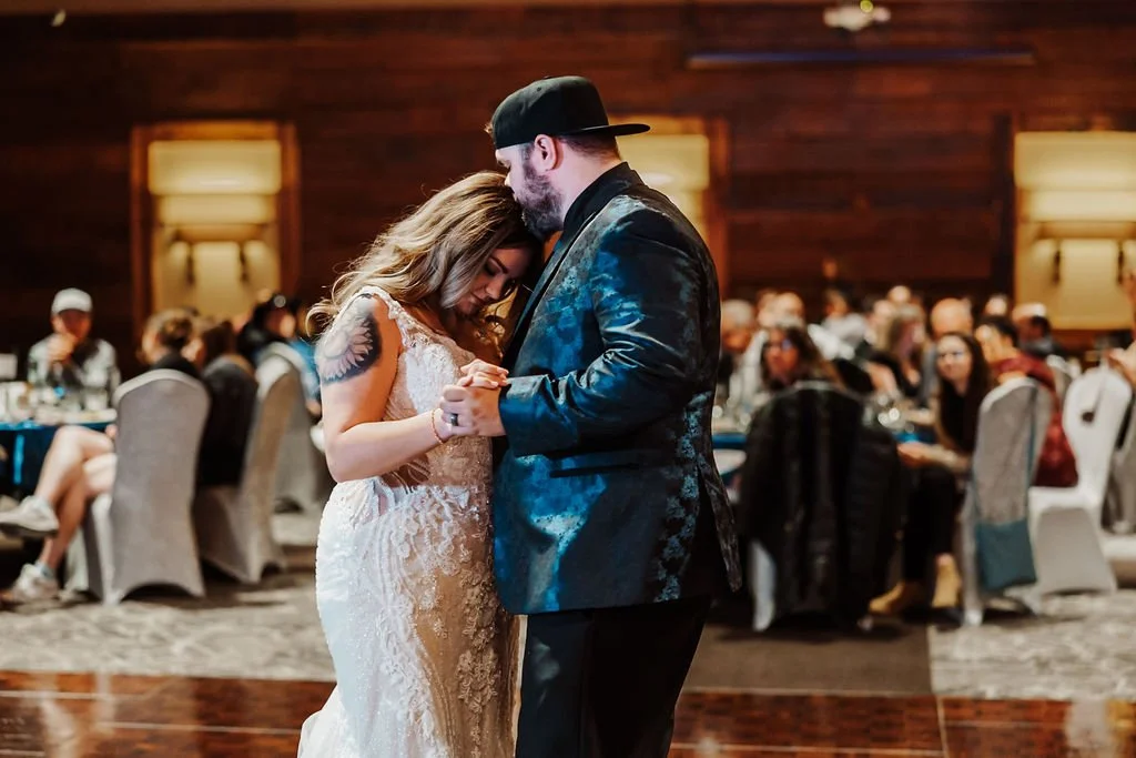 A newlywed couple dances closely at their wedding reception, with guests seated at tables in the background.