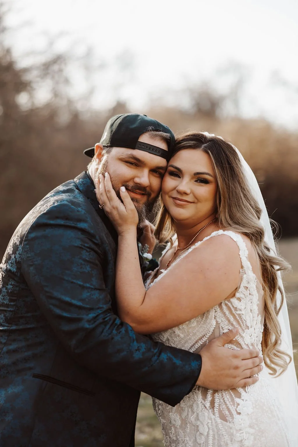 A newlywed couple hugging outdoors, dressed in wedding attire, with the bride in a lace wedding dress and the groom in a dark suit and baseball cap, both smiling.