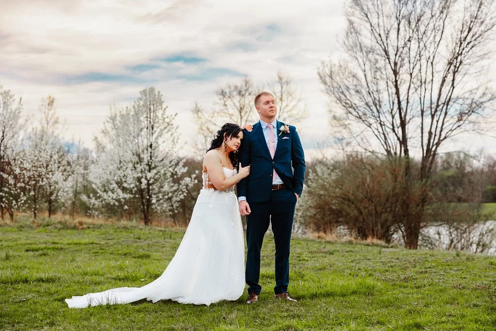 A bride in a white wedding dress and a groom in a navy suit standing outdoors on grass, with trees in bloom and a river in the background during daytime.