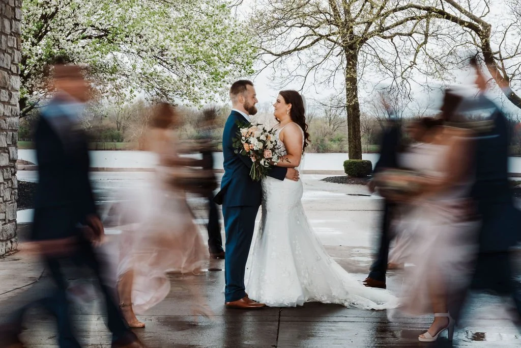 A bride and groom stand facing each other outdoors, holding a bouquet of flowers, surrounded by blurred wedding guests walking past on a rainy day with trees and a body of water in the background.