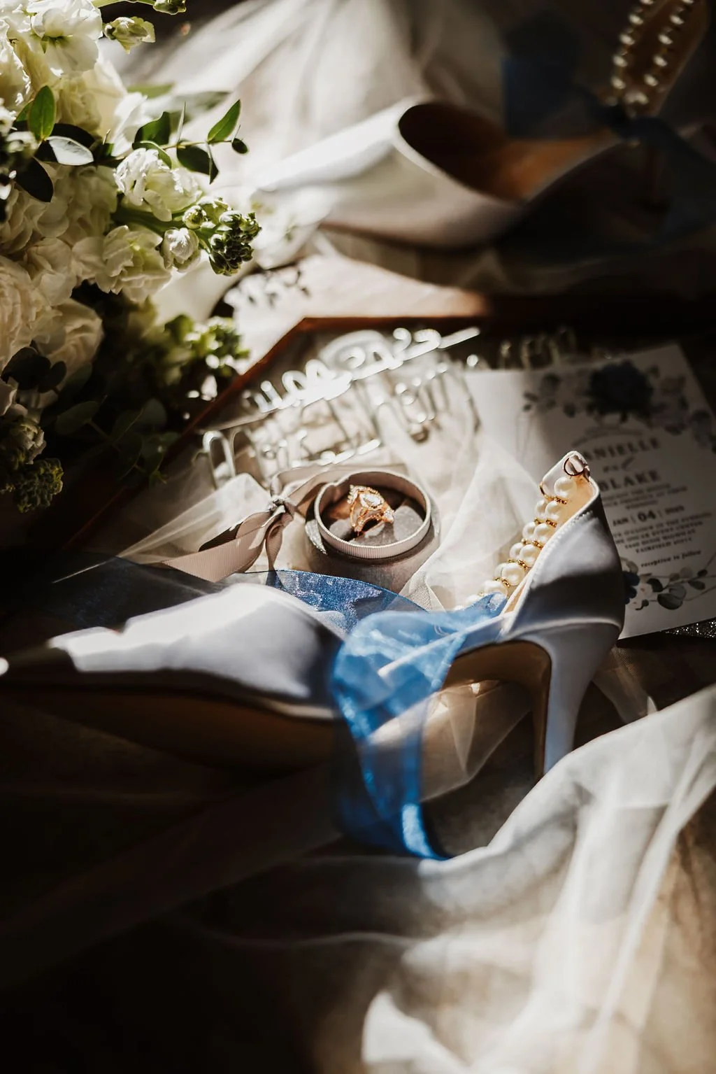 A wedding shoe, jewelry, a wedding invitation, and flowers arranged on a table, with sunlight highlighting the items.