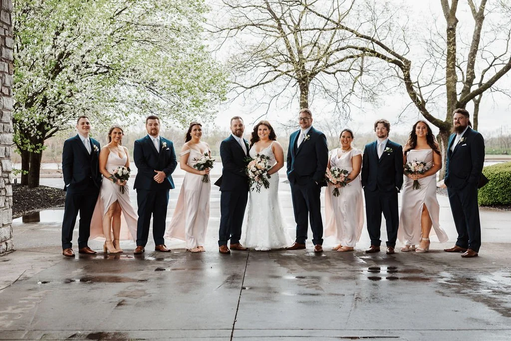Wedding party standing outdoors during rainy weather, dressed in formal attire, with trees and a pond in the background.