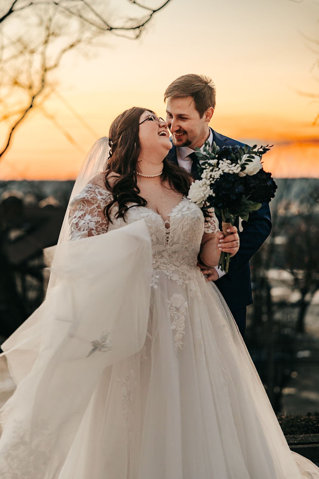 A bride and groom celebrating their wedding at sunset, smiling and looking at each other. The bride wears a lace wedding gown and veil, holding a bouquet of white and dark flowers. The groom wears a suit, embracing the bride.