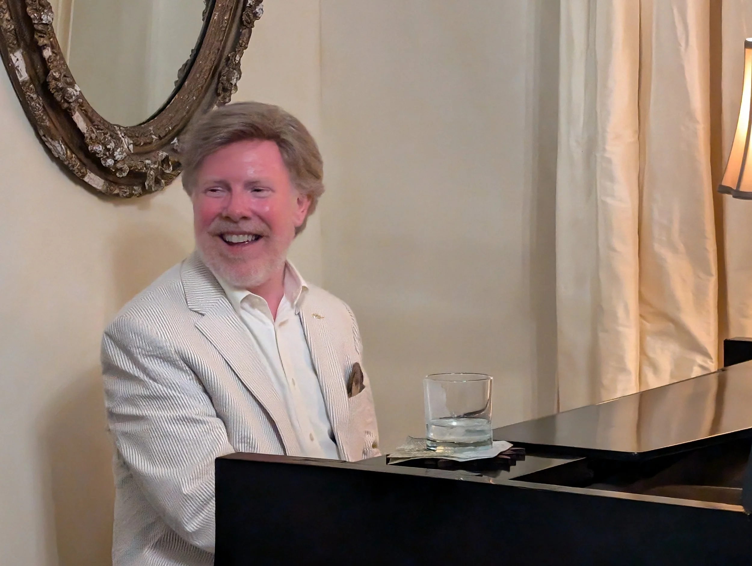 A man with gray hair and a beard wearing a light-colored suit jacket and white shirt, sitting at a black piano with a glass of water on a napkin nearby, smiling and looking to the left. There is a decorative mirror and a lamp in the background.
