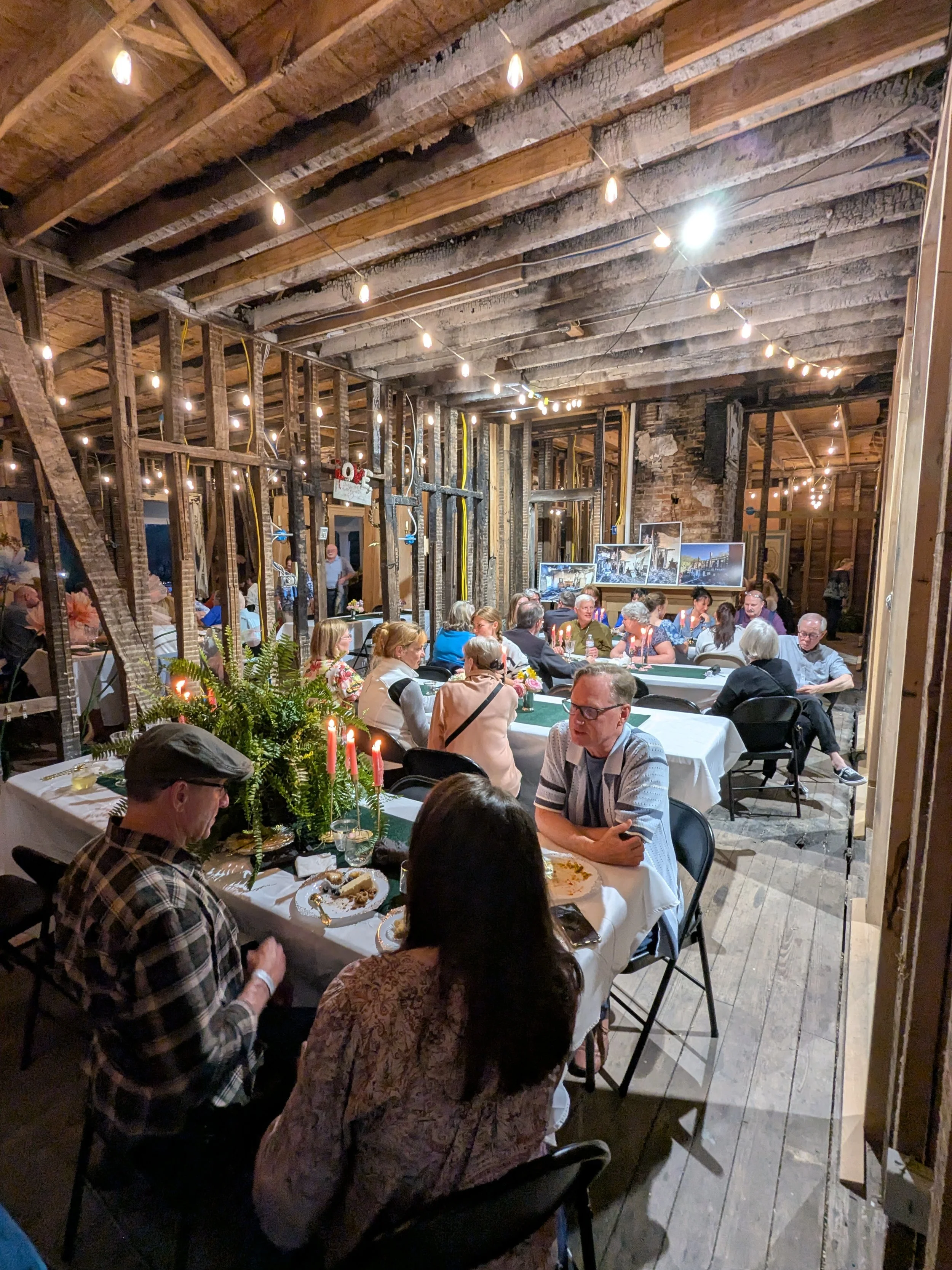 Indoor gathering of people at a rustic banquet with wooden beams, string lights, candles, and framed photos