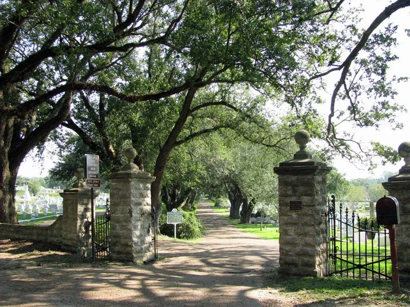 Entrance to a cemetery with stone pillars and wrought iron gates, surrounded by large, leafy trees.
