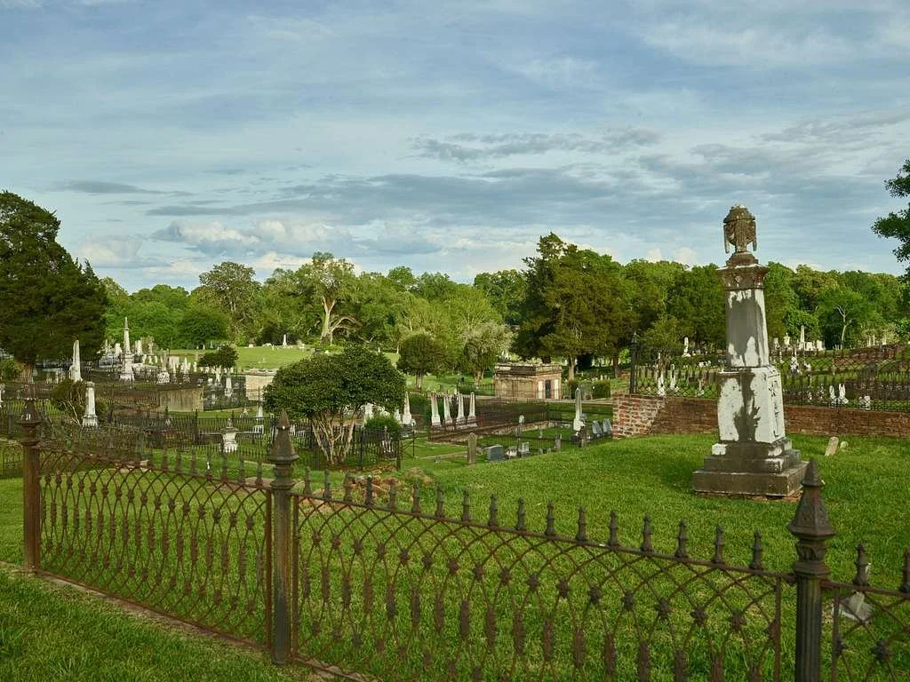 View of a historic cemetery with old white gravestones, statues, and iron fences surrounded by green trees and grass under a partly cloudy sky.