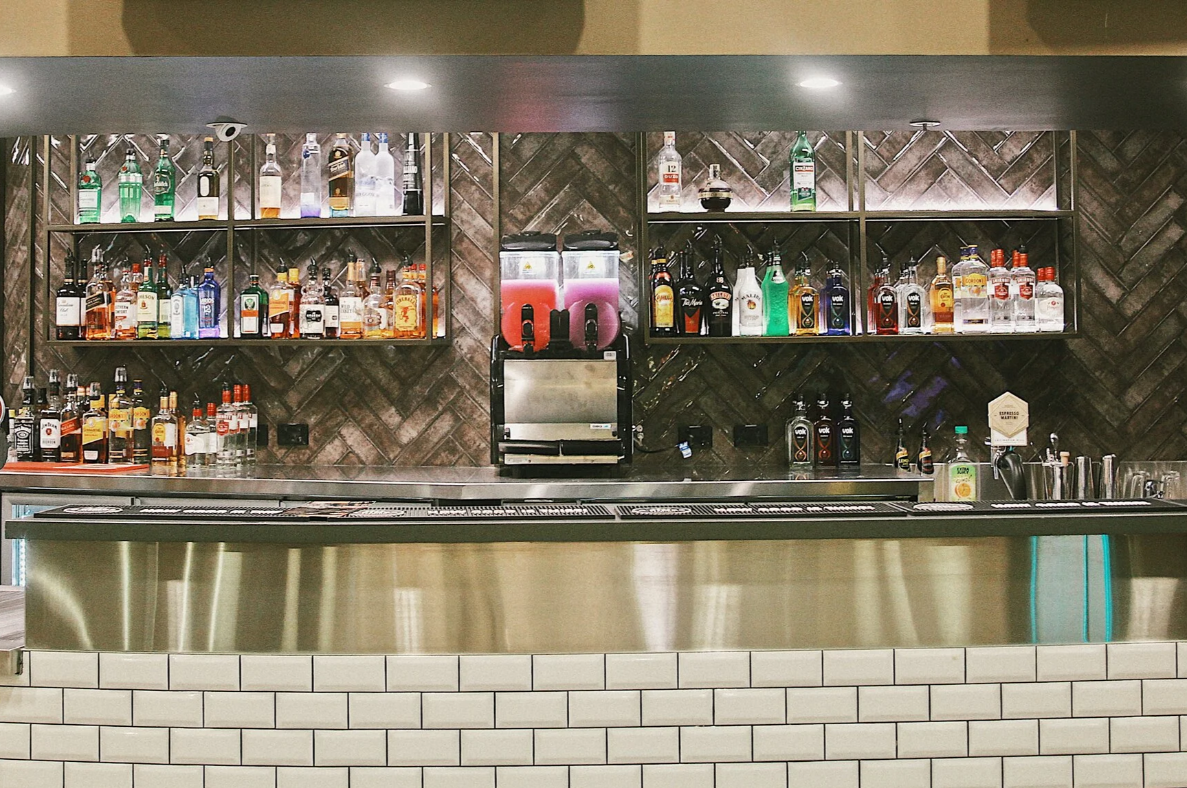 A bar with a variety of liquor bottles on shelves, a beer tap, and a cash register, with a tiled bar counter and brick wall background.
