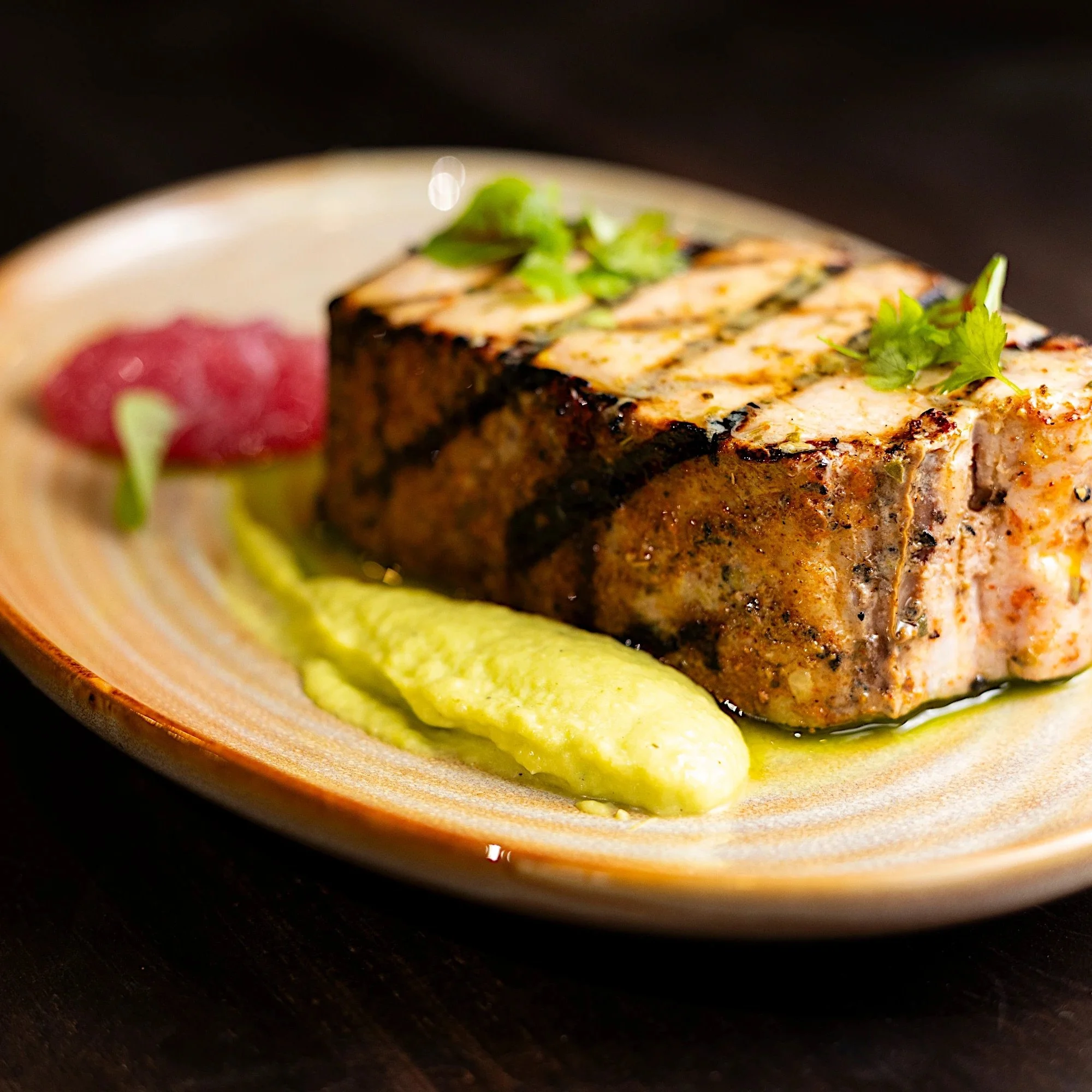 A cooked piece of fish garnished with herbs, served on a plate with green puree, a dollop of red sauce, and microgreens.