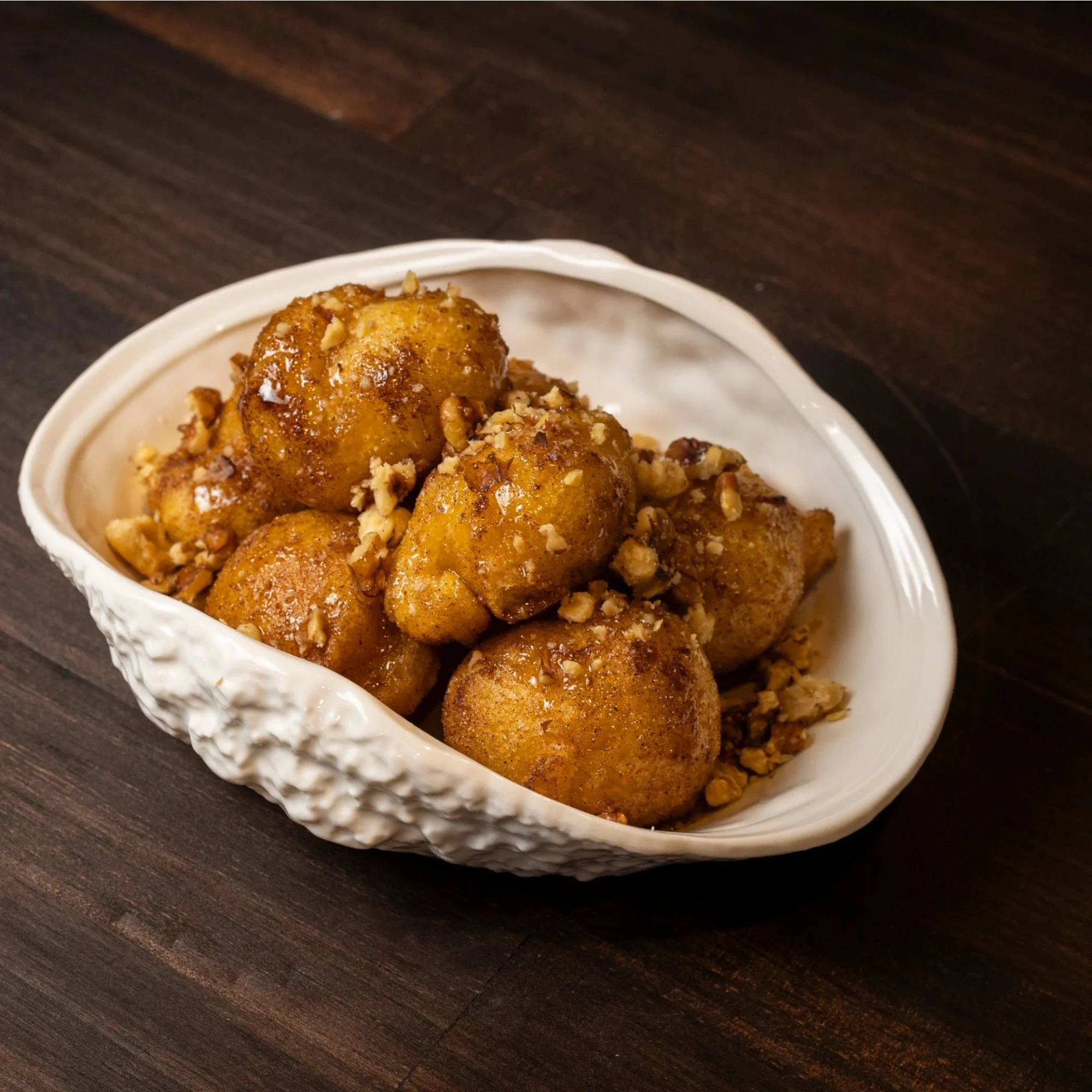A white textured bowl filled with golden brown fried doughnuts covered with crushed nuts on a dark wooden surface.