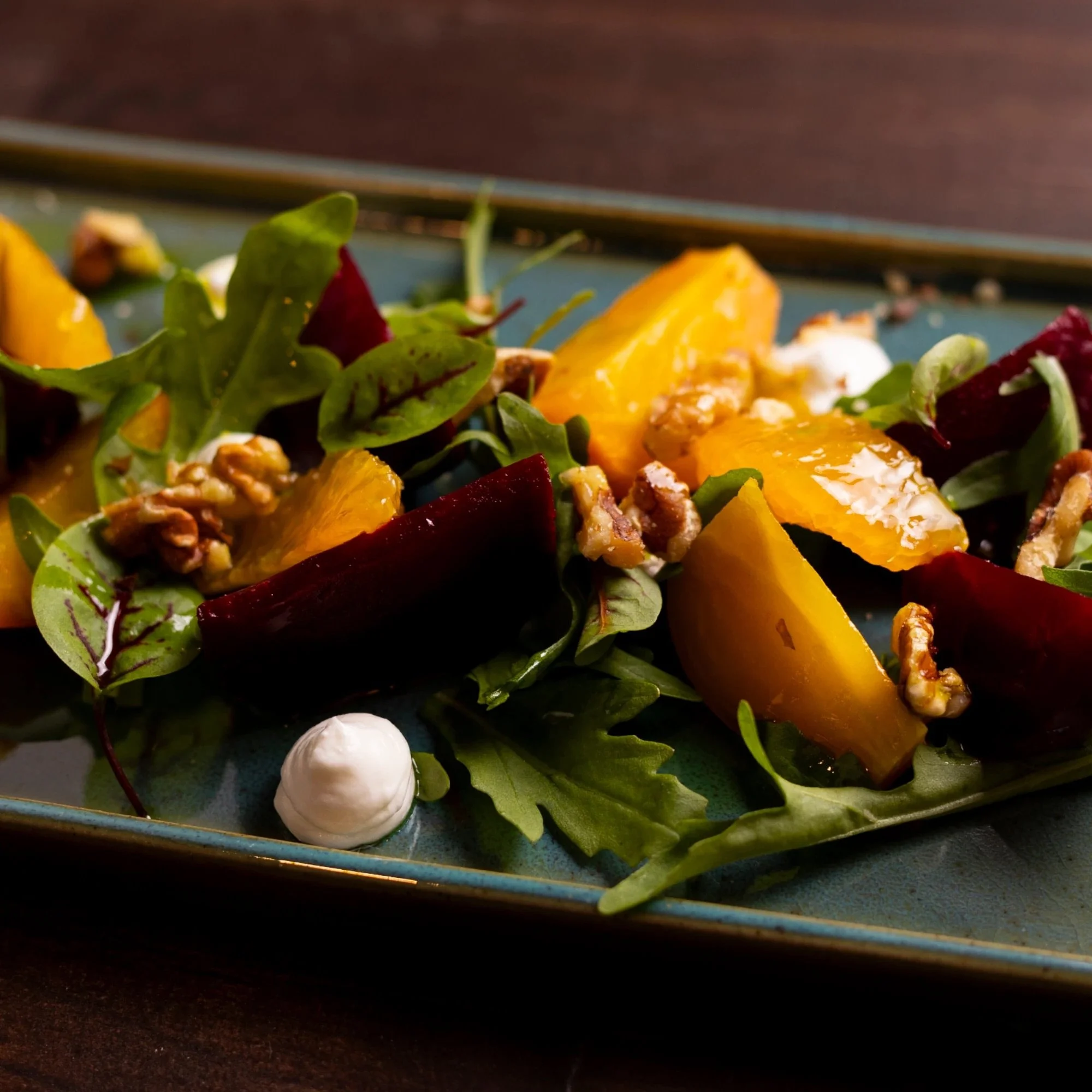 A salad with leafy greens, roasted beets, chunks of yellow fruit, walnuts, and a dollop of white dressing on a dark ceramic plate.