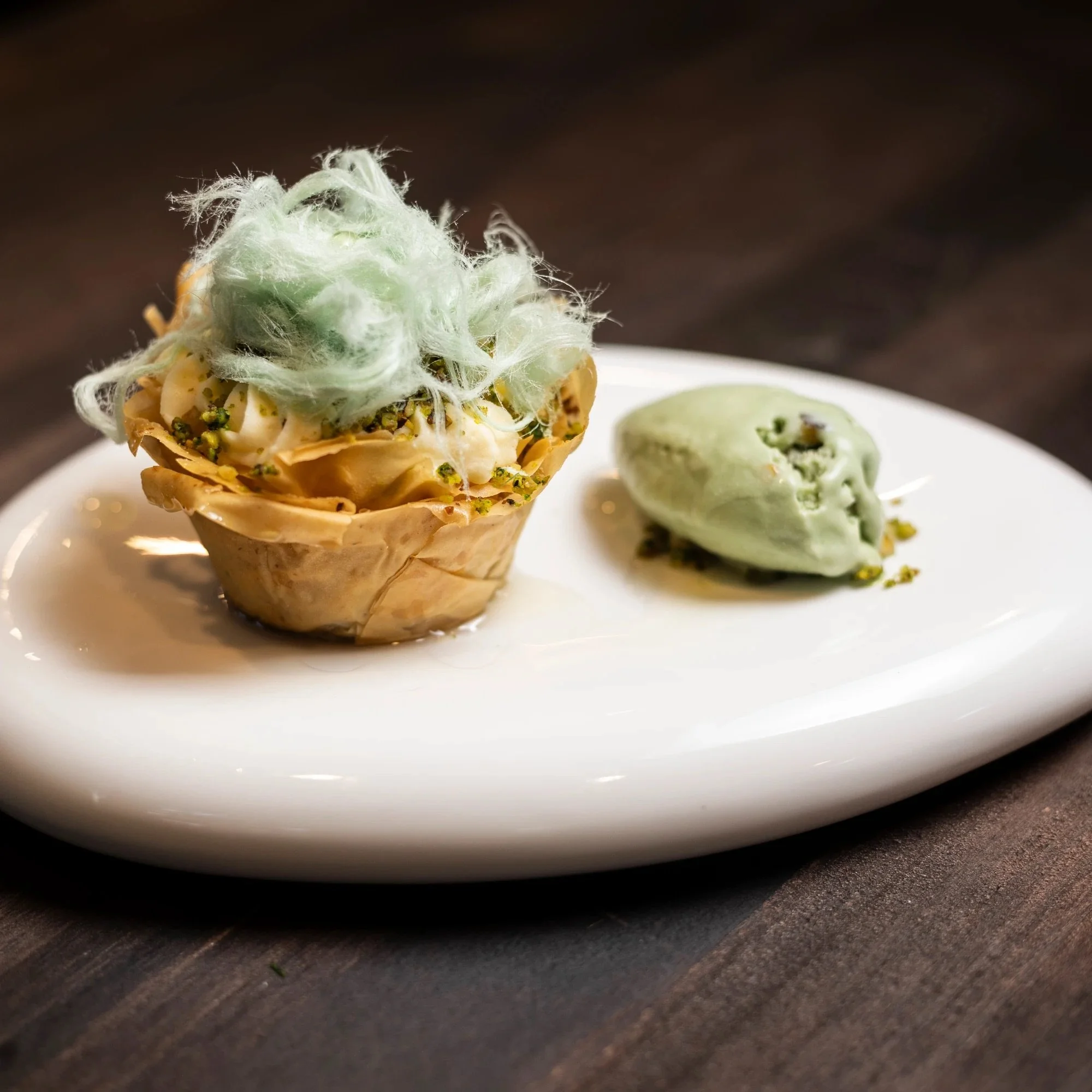 A dessert on a white plate featuring a cone with creamy filling topped with green cotton candy and crushed pistachios, accompanied by a scoop of green ice cream with pistachio pieces.