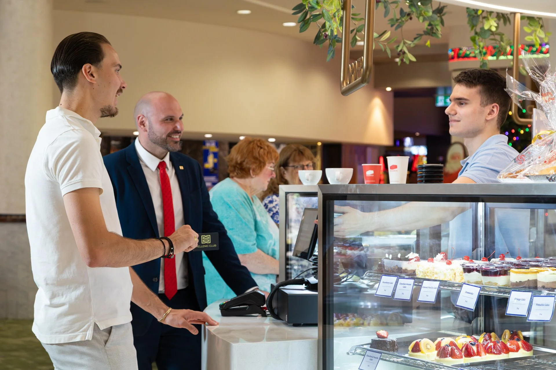 People at a bakery counter ordering desserts, with cakes and pastries on display.