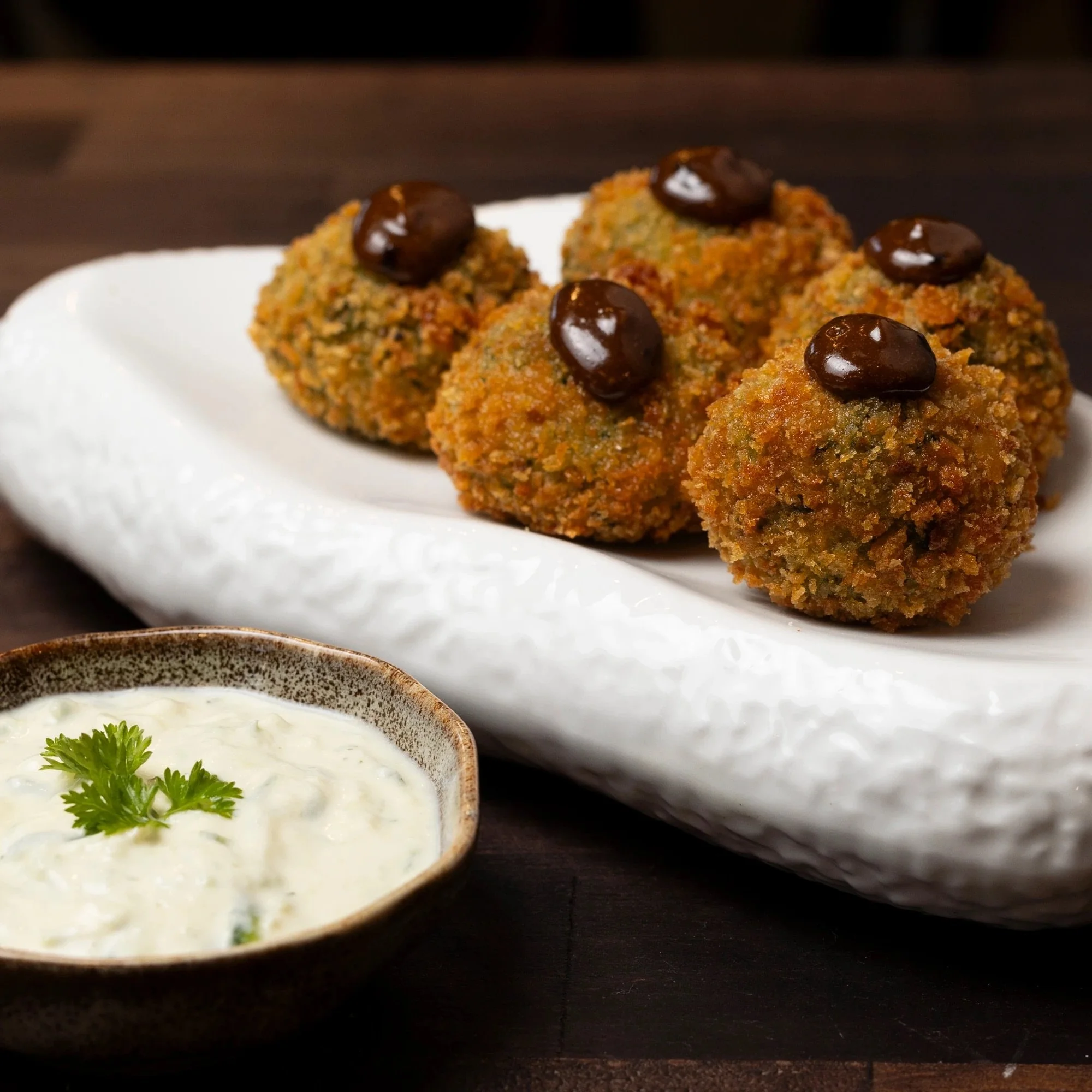 Breaded and fried appetizer balls topped with dark sauce, served with a side of white creamy dipping sauce garnished with a sprig of cilantro on a ceramic plate.