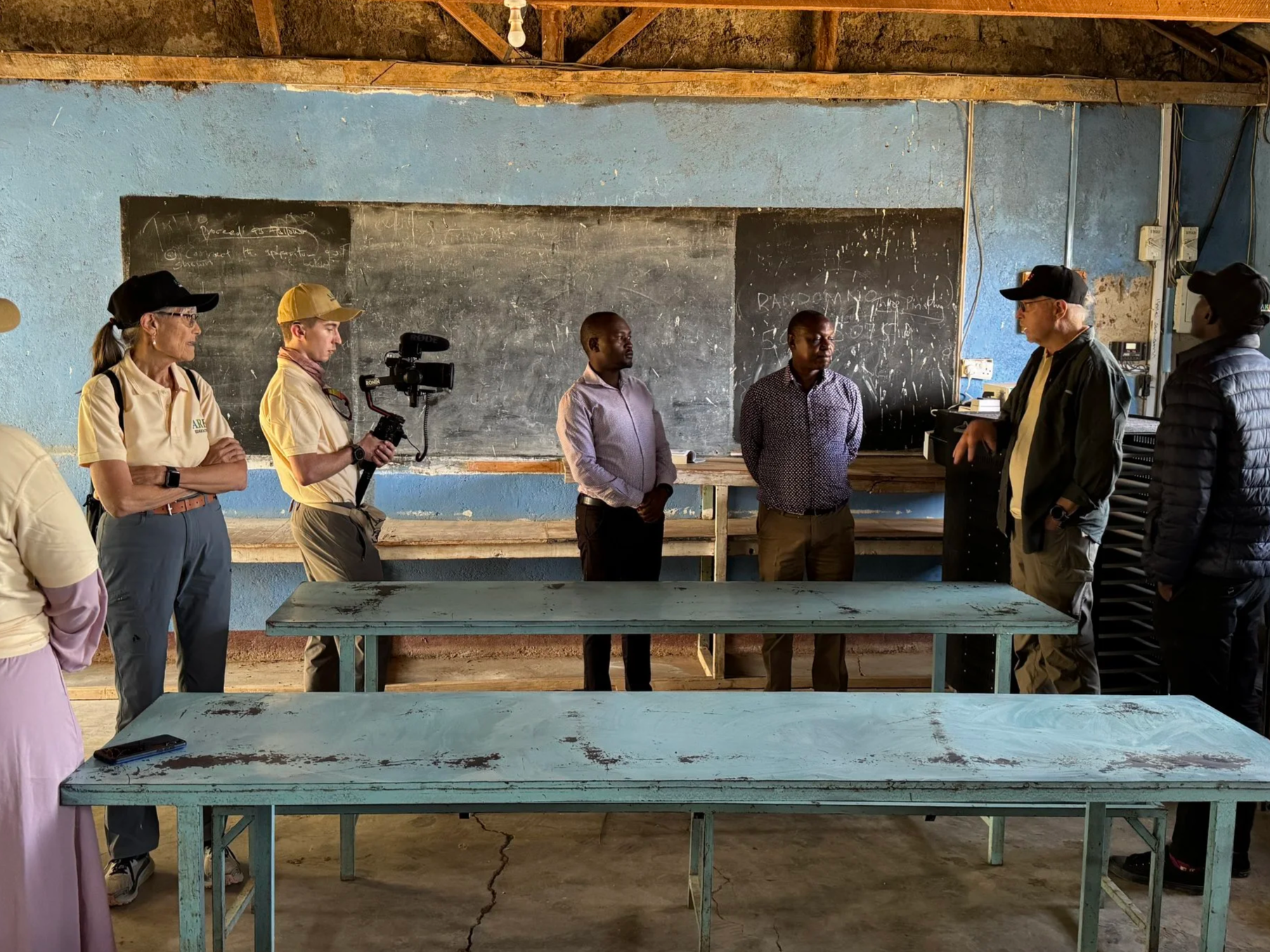 Group of five people standing in a row inside a classroom, facing one another. The room has a blue wall with blackboards, and some papers and electrical outlets are visible. The tables in front are painted light blue and show signs of wear.