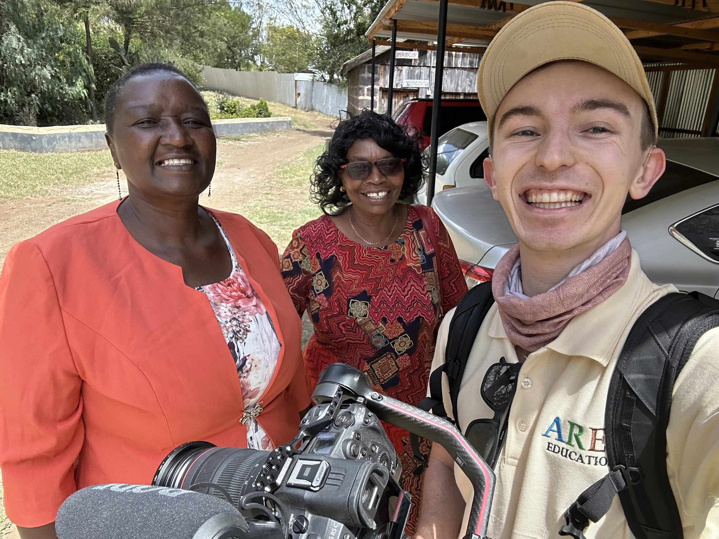 Three smiling women and one smiling man taking a selfie outdoors near parked cars and a wooden shed, with trees and a fence in the background.