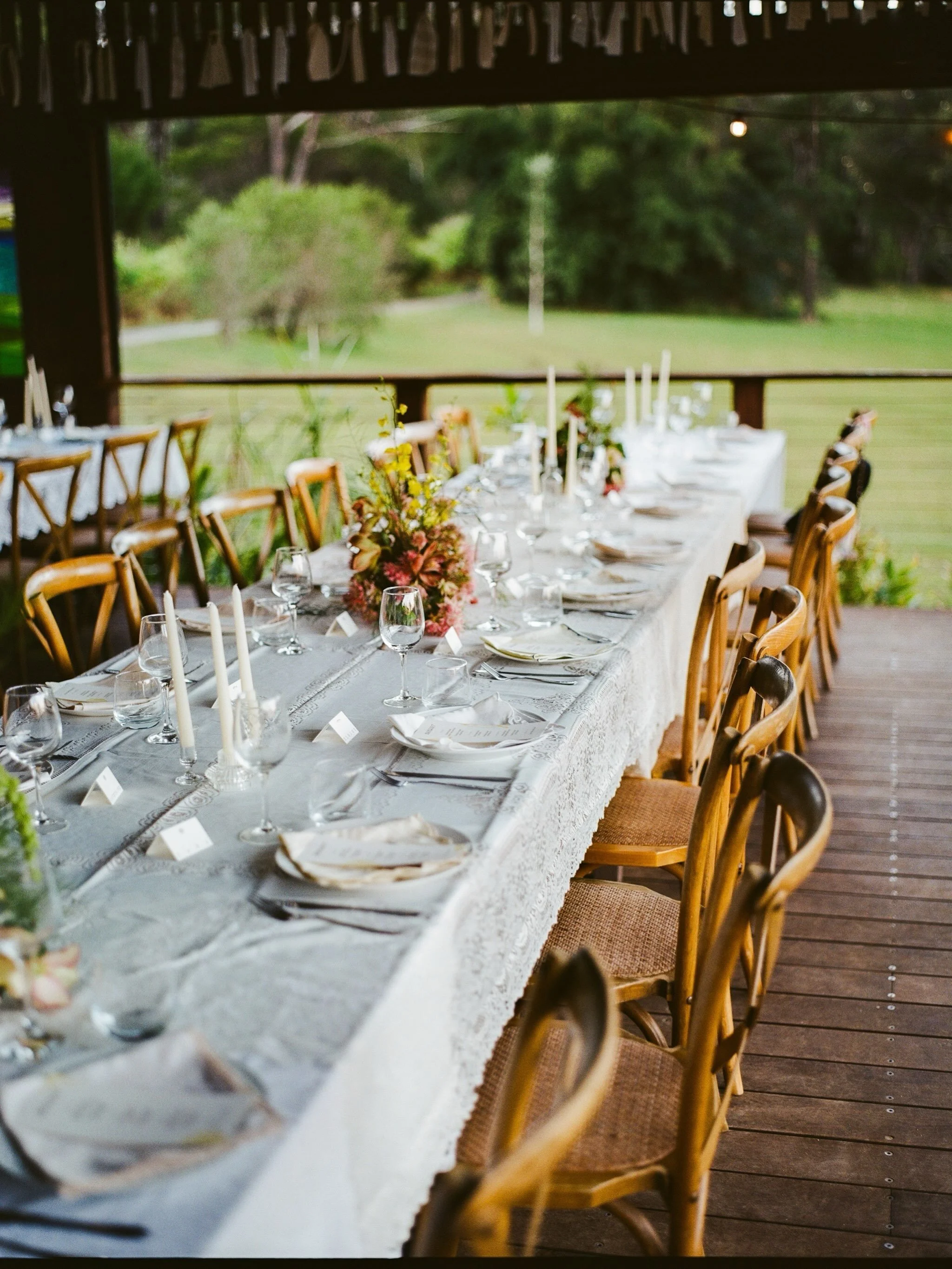 A long dining table set for a meal on a covered porch with wooden chairs, white tablecloth, glassware, candles, and floral centerpieces, overlooking an outdoor green landscape.